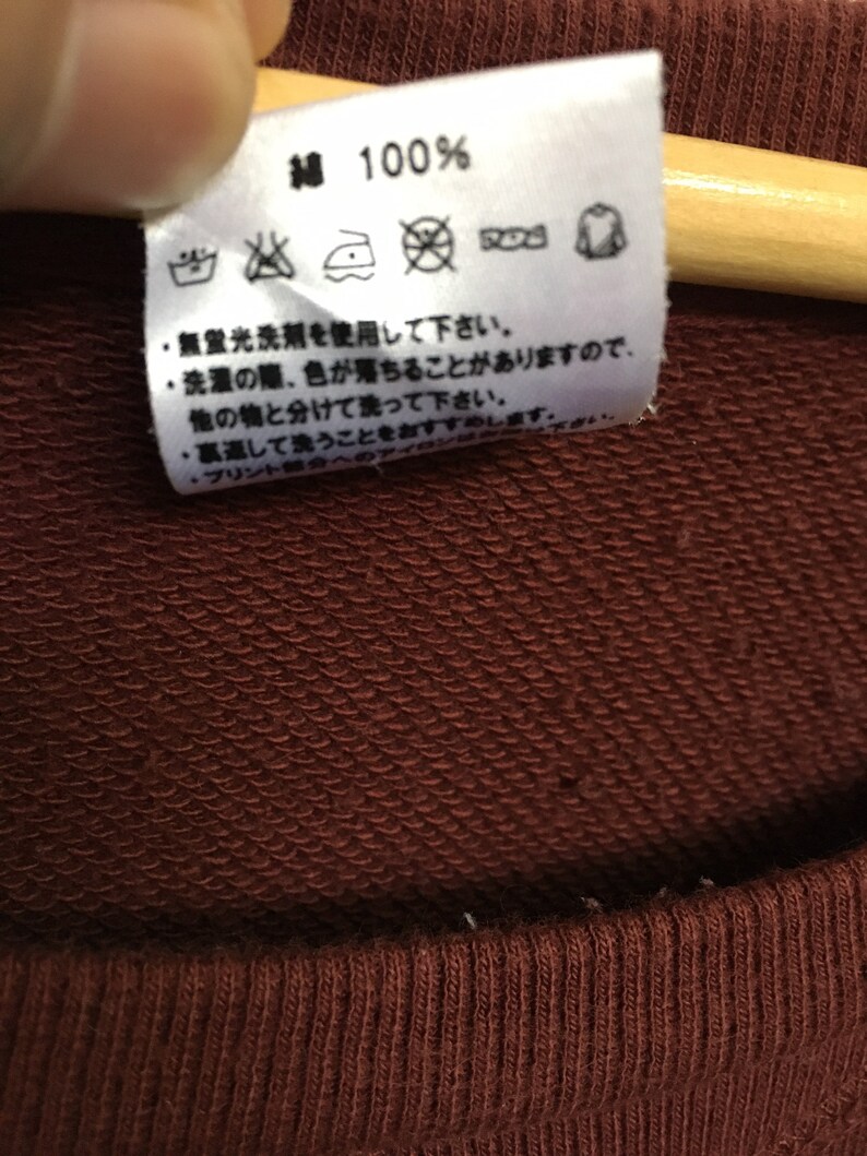 May include: A close-up of a brown care tag with white text and symbols. The tag says "Cotton 100%" and includes instructions for washing, bleaching, ironing, and drying.