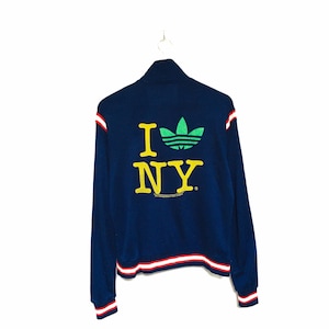 Könnte beinhalten: Eine marineblaue Reißverschlussjacke mit roten und weißen Streifen an den Bündchen und am Bund. Die Rückseite der Jacke zeigt ein gelbes "I ♥ NY"-Motiv mit einem grünen Adidas-Logo.