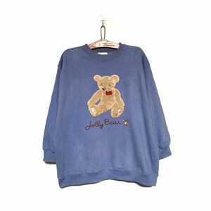May include: A blue sweatshirt with a Jolly Bear design. The bear is tan with a red bow tie. The words "Jolly Bear" are embroidered below the bear. The sweatshirt is hanging on a wooden hanger.
