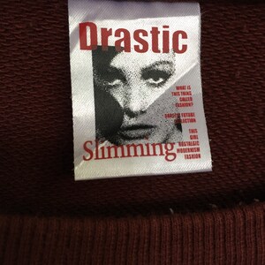 May include: A white tag with the words "Drastic Slimming" in red lettering. The tag features a black and white image of a woman's face with the text "What is this thing called fashion? Drastic Future Collection This Girl Nostalgic Modernism Fashion".