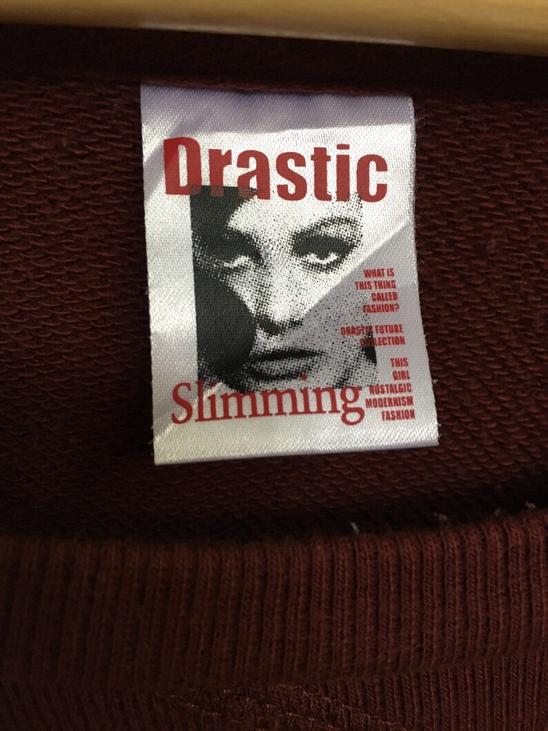 May include: A white tag with the words "Drastic Slimming" in red lettering. The tag features a black and white image of a woman's face with the text "What is this thing called fashion? Drastic Future Collection This Girl Nostalgic Modernism Fashion".