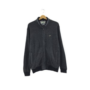 Könnte beinhalten: Eine dunkelgraue Reißverschlussjacke mit geripptem Kragen und Bündchen. Die Jacke hat ein kleines Logo auf der linken Brust und zwei Taschen an den Seiten.