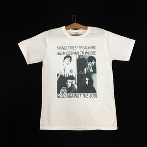 Puede incluir: Camiseta blanca con una foto en blanco y negro de la banda Manic Street Preachers y el título del álbum "From Despair to Where Gold Against the Soul."