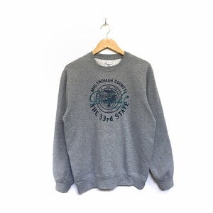 May include: A heather gray sweatshirt with a crew neck and long sleeves. The front features a teal and white graphic with the text "Multnomah County, The 33rd State."