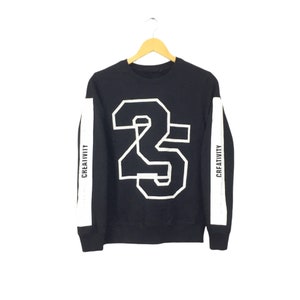 Puede incluir: Sudadera negra con un gran número blanco 23 en la parte delantera. Rayas blancas con la palabra "CREATIVITY" recorren las mangas. La sudadera está colgada de una percha de madera.
