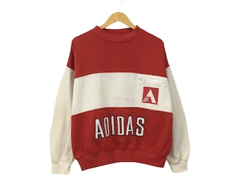 May include: Red, white, and cream colored sweatshirt with a large "ADIDAS" logo on the front. The sweatshirt has a white stripe across the chest and a small pocket with a red triangle logo on the right side.