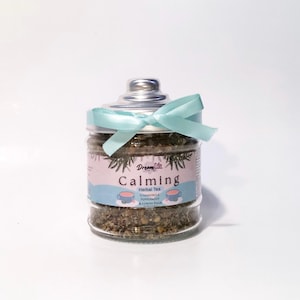 Op de afbeelding: Een glazen pot gevuld met kruidenthee, met het label "Calming". De pot heeft een zilveren deksel en is vastgebonden met een lichtblauw lint. Het label vermeldt kamille, pepermunt en citroenmelisse.