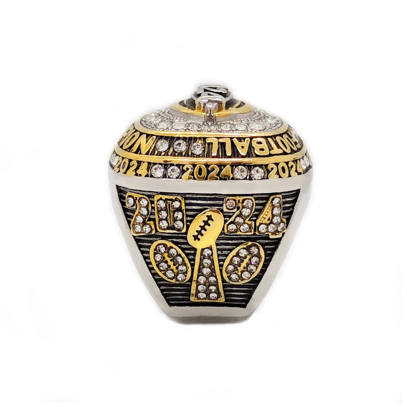 May include: A gold and silver football championship ring with the year 2024 displayed. The ring features the words "FOOTBALL WIN" and is adorned with numerous clear gemstones. The design includes a football and the number 24.