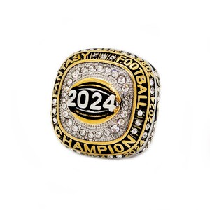 May include: A gold-toned ring with a square face, encrusted with clear gemstones. The face features a football design with the year "2024" and the words "FANTASY FOOTBALL CHAMPION" in black and gold lettering. The ring is set against a white background.