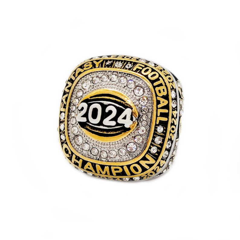 May include: A gold-toned ring with a square face, encrusted with clear gemstones. The face features a football design with the year "2024" and the words "FANTASY FOOTBALL CHAMPION" in black and gold lettering. The ring is set against a white background.