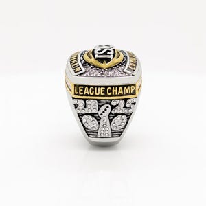 Puede incluir: Un anillo de campeonato plateado y dorado con el texto "LEAGUE CHAMP" y el a&ntilde;o "2022". El anillo tiene un dise&ntilde;o de escudo con las letras "IS" en la parte superior. El anillo est&aacute; sobre un fondo blanco.