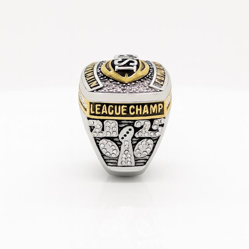Puede incluir: Un anillo de campeonato plateado y dorado con el texto "LEAGUE CHAMP" y el a&ntilde;o "2022". El anillo tiene un dise&ntilde;o de escudo con las letras "IS" en la parte superior. El anillo est&aacute; sobre un fondo blanco.