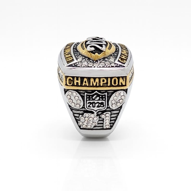 Puede incluir: Un anillo de campeonato plateado y dorado con la palabra "CHAMPION" en letras doradas. El anillo presenta un dise&ntilde;o con el a&ntilde;o "2023" y est&aacute; adornado con peque&ntilde;as piedras transparentes. El anillo est&aacute; sobre un fondo blanco.