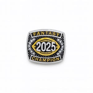 Puede incluir: Anillo plateado con una cara rectangular. El anillo presenta las palabras "FANTASY CHAMPION" y el n&uacute;mero "2025" en oro y blanco. Peque&ntilde;as piedras transparentes rodean el dise&ntilde;o central.