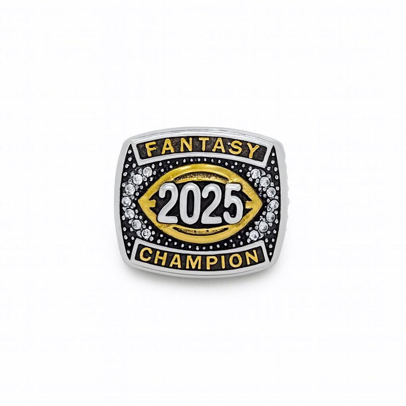 Puede incluir: Anillo plateado con una cara rectangular. El anillo presenta las palabras "FANTASY CHAMPION" y el n&uacute;mero "2025" en oro y blanco. Peque&ntilde;as piedras transparentes rodean el dise&ntilde;o central.