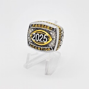 Puede incluir: Un anillo de campeonato de f&uacute;tbol de fantas&iacute;a plateado y dorado. El anillo presenta el a&ntilde;o "2025" en el centro, rodeado por las palabras "FANTASY CHAMPION" y "LEAGUE CHAMP". El anillo est&aacute; incrustado con peque&ntilde;as piedras transparentes.