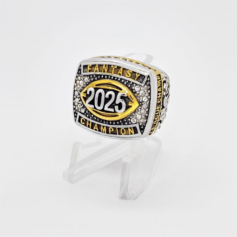 Puede incluir: Un anillo de campeonato de f&uacute;tbol de fantas&iacute;a plateado y dorado. El anillo presenta el a&ntilde;o "2025" en el centro, rodeado por las palabras "FANTASY CHAMPION" y "LEAGUE CHAMP". El anillo est&aacute; incrustado con peque&ntilde;as piedras transparentes.