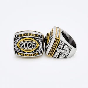 Puede incluir: Dos anillos de campeonato de f&uacute;tbol fantasy plateados y dorados. Uno muestra "2025" en el centro, rodeado de "FANTASY" y "CHAMPION". El otro anillo tiene "LEAGUE CHAMP" en el lateral y est&aacute; incrustado con peque&ntilde;as piedras transparentes.