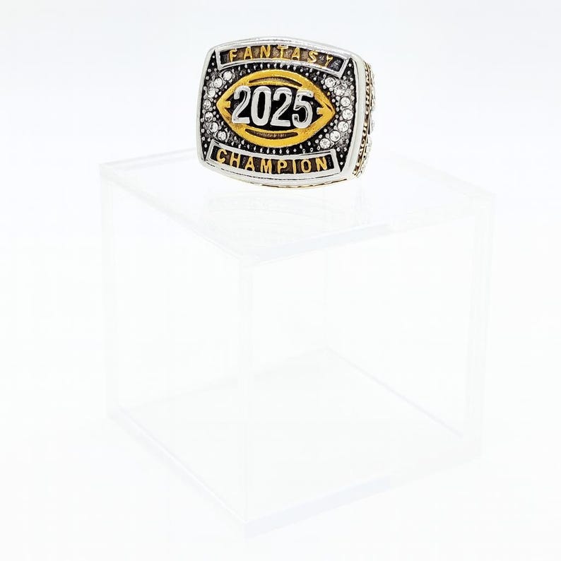 Puede incluir: Un anillo plateado y dorado con el texto "FANTASY CHAMPION" y "2025". El anillo tiene una cara rectangular con un dise&ntilde;o en forma de bal&oacute;n de f&uacute;tbol en el centro. El anillo est&aacute; incrustado con peque&ntilde;as piedras transparentes.