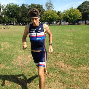 May include: A man wearing a blue and black athletic shirt with the text "Aquaman.aus" and a blue headband. He is running on a grassy field.