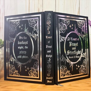 Puede incluir: Un libro negro con detalles en l&aacute;mina plateada. La portada presenta dise&ntilde;os florales, estrellas y el t&iacute;tulo "A Court of Frost and Starlight" con el nombre del autor. El lomo tambi&eacute;n tiene el t&iacute;tulo. El libro est&aacute; abierto, revelando las p&aacute;ginas interiores.