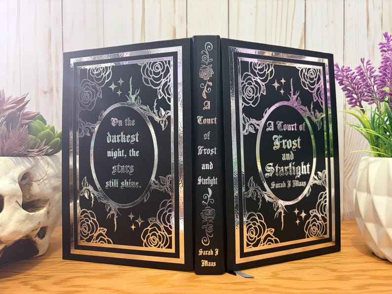 Puede incluir: Un libro negro con detalles en l&aacute;mina plateada. La portada presenta dise&ntilde;os florales, estrellas y el t&iacute;tulo "A Court of Frost and Starlight" con el nombre del autor. El lomo tambi&eacute;n tiene el t&iacute;tulo. El libro est&aacute; abierto, revelando las p&aacute;ginas interiores.