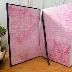 May include: Open journal with pink pages featuring a castle illustration and script. The journal has a black spine and a silver ribbon bookmark. The pages are open, revealing the interior design. The journal is on a wooden surface.