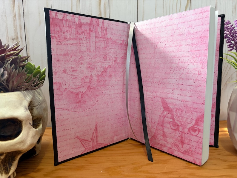 May include: Open journal with pink pages featuring a castle illustration and script. The journal has a black spine and a silver ribbon bookmark. The pages are open, revealing the interior design. The journal is on a wooden surface.