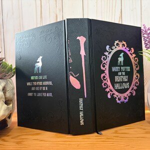 May include: Black book with the title "Harry Potter and the Deathly Hallows" in a decorative frame. The spine features a wand illustration. The cover has a deer graphic and text: "Neither can live while the other survives, and one of us is about to live for good."