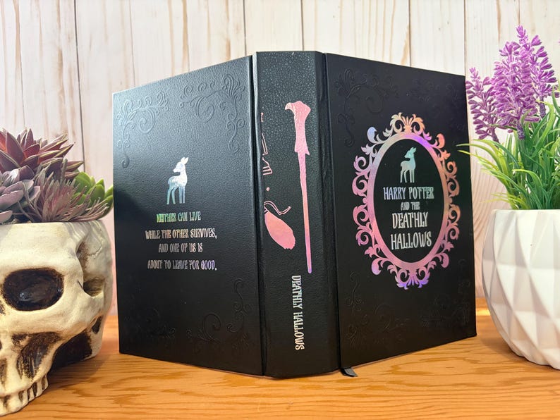 May include: Black book with the title "Harry Potter and the Deathly Hallows" in a decorative frame. The spine features a wand illustration. The cover has a deer graphic and text: "Neither can live while the other survives, and one of us is about to live for good."