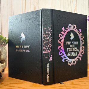 May include: Black book with the title "Harry Potter and the Prisoner of Azkaban" in a decorative frame. The book has a unicorn silhouette and the text "BAR TO HE YOU WON'T GO LOOKING FOR BLACK". The spine features a cactus illustration.