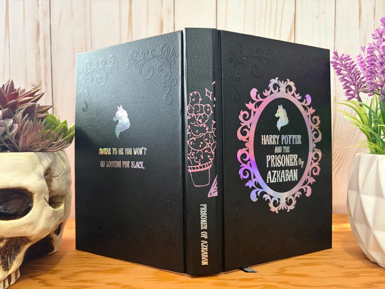 May include: Black book with the title "Harry Potter and the Prisoner of Azkaban" in a decorative frame. The book has a unicorn silhouette and the text "BAR TO HE YOU WON'T GO LOOKING FOR BLACK". The spine features a cactus illustration.