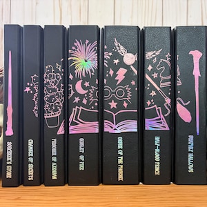 May include: A collection of seven black books with holographic pink and purple illustrations and titles. The titles include "Sorcerer's Stone", "Chamber of Secrets", and "Deathly Hallows". The books are arranged on a wooden surface.