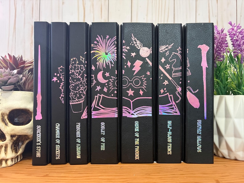 May include: A collection of seven black books with holographic pink and purple illustrations and titles. The titles include "Sorcerer's Stone", "Chamber of Secrets", and "Deathly Hallows". The books are arranged on a wooden surface.