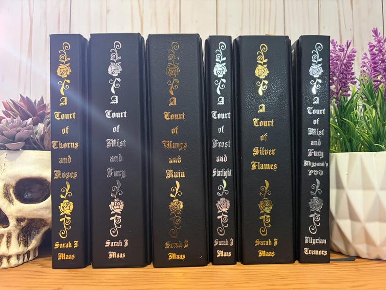 May include: A collection of six black hardcover books with gold, silver, and pink floral designs. The titles include "A Court of Thorns and Roses," "A Court of Mist and Fury," and "A Court of Wings and Ruin."