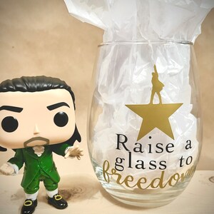 K&ouml;nnte beinhalten: Eine gr&uuml;ne und schwarze Popfigur eines Mannes mit wei&szlig;em Kragen und einem goldenen Stern auf einem Weinglas, auf dem "Raise a glass to freedom" steht.