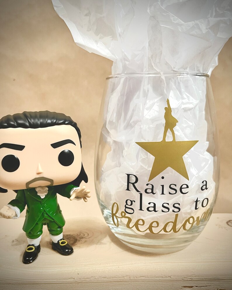 K&ouml;nnte beinhalten: Eine gr&uuml;ne und schwarze Popfigur eines Mannes mit wei&szlig;em Kragen und einem goldenen Stern auf einem Weinglas, auf dem "Raise a glass to freedom" steht.