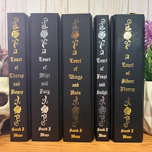 Puede incluir: Una colecci&oacute;n de cinco libros de tapa dura negros con dise&ntilde;os florales dorados y plateados. Los t&iacute;tulos incluyen "A Court of Thorns and Roses", "A Court of Mist and Fury", "A Court of Wings and Ruin", "A Court of Frost and Starlight" y "A Court of Silver Flames".
