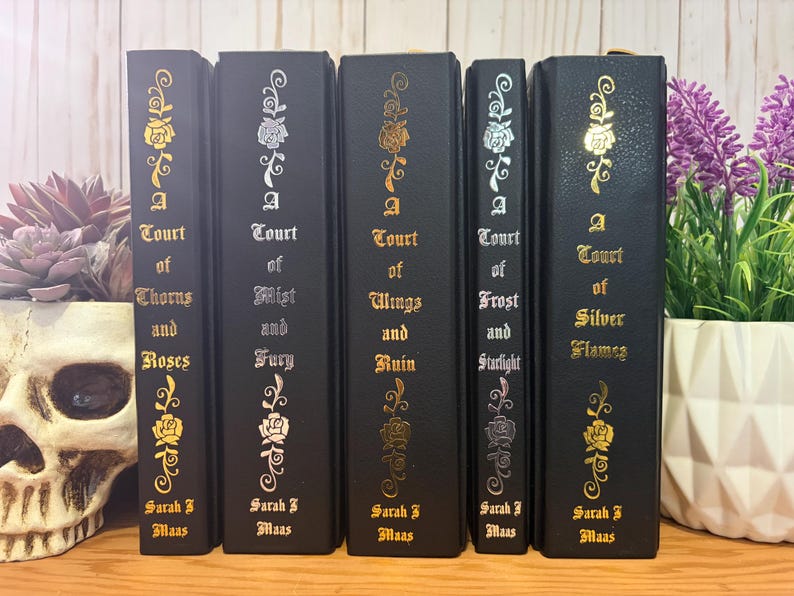 Puede incluir: Una colecci&oacute;n de cinco libros de tapa dura negros con dise&ntilde;os florales dorados y plateados. Los t&iacute;tulos incluyen "A Court of Thorns and Roses", "A Court of Mist and Fury", "A Court of Wings and Ruin", "A Court of Frost and Starlight" y "A Court of Silver Flames".
