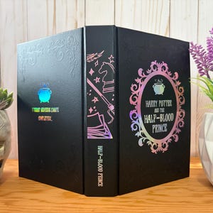 May include: A black book with the title "Harry Potter and the Half-Blood Prince" in a decorative frame. The book has holographic accents and the text "I TRUST SEPADS SWATE" on the spine. The book is on a wooden surface.