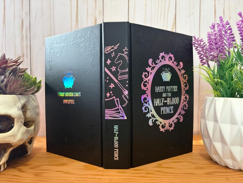 May include: A black book with the title "Harry Potter and the Half-Blood Prince" in a decorative frame. The book has holographic accents and the text "I TRUST SEPADS SWATE" on the spine. The book is on a wooden surface.