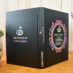 May include: Black book with embossed floral design and the text "Harry Potter and the Chamber of Secrets." The cover features a snake emblem and the quote "Harry Potter must not go back to Hogwarts." The book is displayed on a wooden surface.