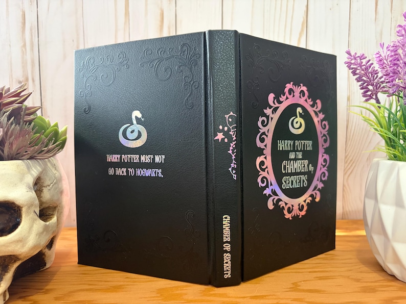 May include: Black book with embossed floral design and the text "Harry Potter and the Chamber of Secrets." The cover features a snake emblem and the quote "Harry Potter must not go back to Hogwarts." The book is displayed on a wooden surface.