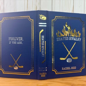 May include: A hardcover book with a navy blue cover and gold foil accents. The title "Heated Rivalry" is displayed with hockey-themed graphics. The author's name, Rachel Reid, is also visible.