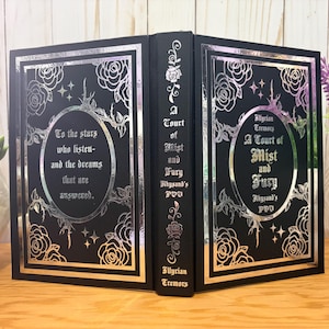 Puede incluir: Un libro negro con detalles en l&aacute;mina plateada. La portada presenta dise&ntilde;os florales, estrellas y el texto "A Court of Mist and Fury" y "To the stars who listen and the dreams that are answered." El lomo tambi&eacute;n tiene texto.