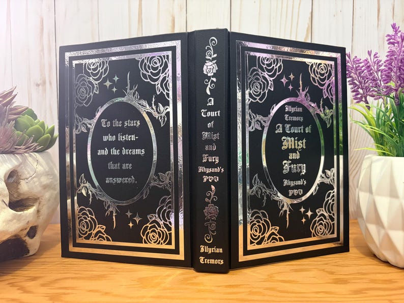 Puede incluir: Un libro negro con detalles en l&aacute;mina plateada. La portada presenta dise&ntilde;os florales, estrellas y el texto "A Court of Mist and Fury" y "To the stars who listen and the dreams that are answered." El lomo tambi&eacute;n tiene texto.