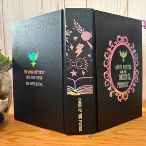 May include: Black book with embossed details and holographic accents. The cover features the text "The world isn't split into good people and death eaters." The spine reads "Order of the Phoenix" with iconic imagery. The book is a collectible item.