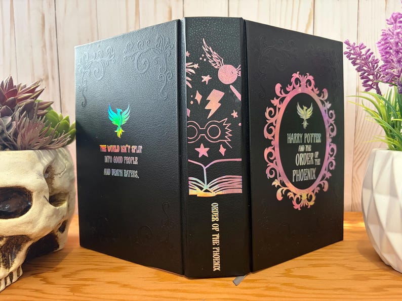May include: Black book with embossed details and holographic accents. The cover features the text "The world isn't split into good people and death eaters." The spine reads "Order of the Phoenix" with iconic imagery. The book is a collectible item.