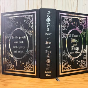 Puede incluir: Libro negro con dise&ntilde;os florales y de estrellas plateadas. El t&iacute;tulo del libro, "A Court of Mist and Fury" de Sarah J. Maas, se muestra en la portada. El lomo tambi&eacute;n presenta el t&iacute;tulo y el nombre del autor. La portada tiene la cita: "To the people who look to the stars and wish."