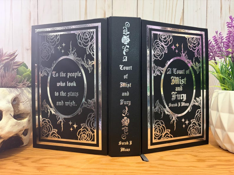 Puede incluir: Libro negro con dise&ntilde;os florales y de estrellas plateadas. El t&iacute;tulo del libro, "A Court of Mist and Fury" de Sarah J. Maas, se muestra en la portada. El lomo tambi&eacute;n presenta el t&iacute;tulo y el nombre del autor. La portada tiene la cita: "To the people who look to the stars and wish."